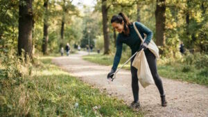 Plogging: Neuer Lauftrend der Joggen und Müllsammeln verbindet 6 Das Bild zeigt eine Frau beim Plogging in einem Stadtpark. Sie hebt mit einer Greifzange Müll vom Boden auf und hat eine Tasche zum Sammeln um die Schulter hängen.