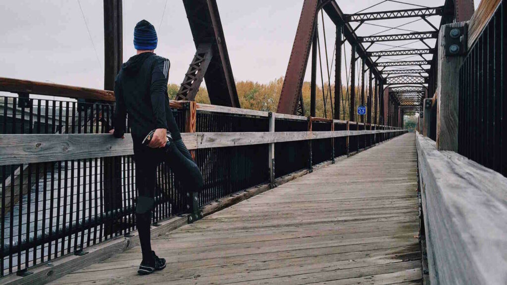 Hyrox: Der trendige Fitness-Wettkampf verbindet Kraftsport mit Laufen und Functional Fitness 3 Das Bild zeigt einen Läufer von hinten auf einer Brücke beim Training für einen Hyrox-Wettkampf. Er macht Dehnübungen für die Beinmuskulatur.