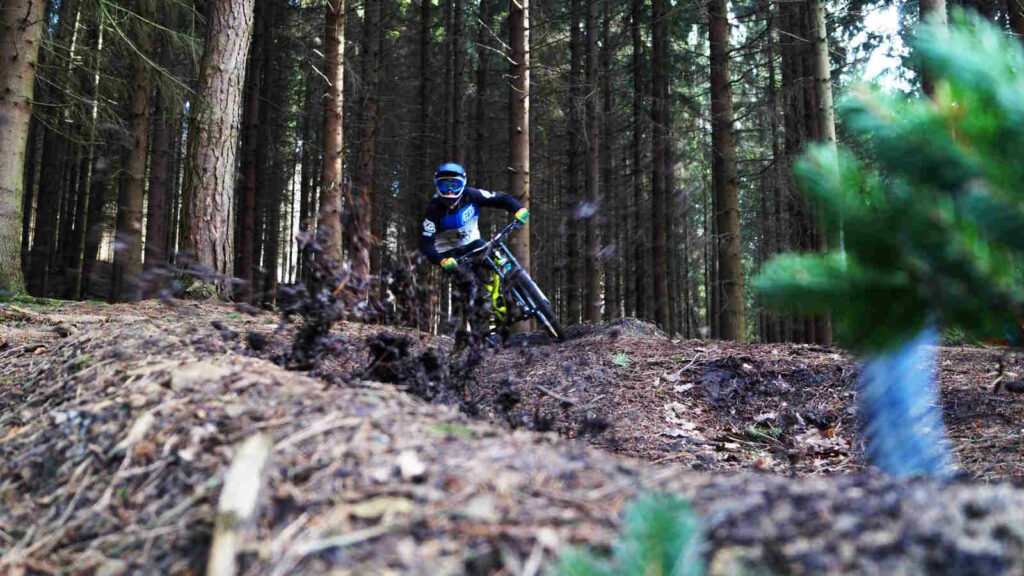 Downhill-Mountainbiking: Technik, Bikes & Tipps für Einsteiger 3 Das Bild zeigt einen Downhill-MTB Trail in einem Wald und einen Fahrer während der Abfahrt.